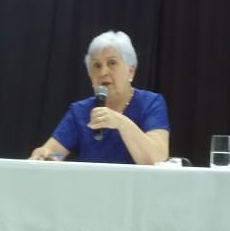 Professora Mirza, sentada, de blusa azul e segurando o microfone